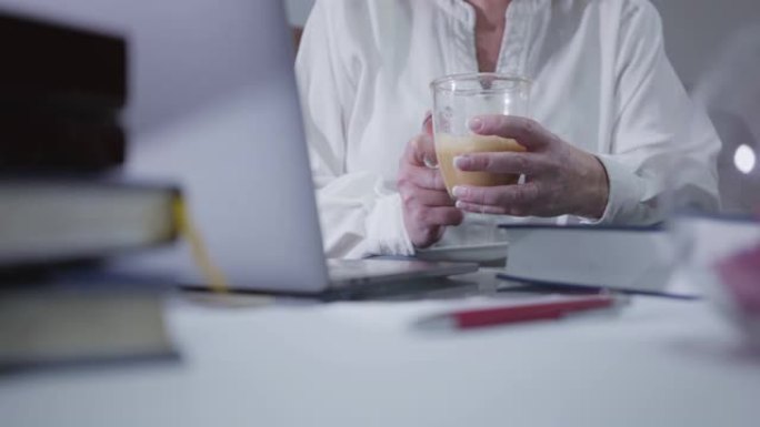 成熟女性手握咖啡杯和使用笔记本电脑的特写镜头。高级白人妇女在家上网。爱好，消遣，休闲，社交网络。