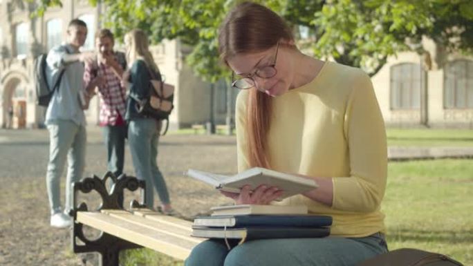 全神贯注的红发女孩在眼镜上看书，模糊的同伴在后台嘲笑她。同伴在大学校园里欺负害羞的年轻白人学生。
