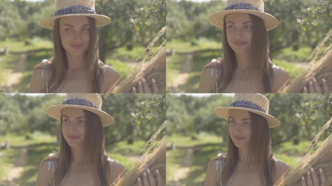 戴着草帽和白色长裙的可爱年轻女子站在绿色的夏日花园里，手里拿着一束干草。