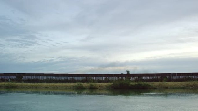 在日落时分的阴天，水沿着一条平行于墨西哥和美国之间的钢板条边界墙 (在美国一侧) 的运河流下