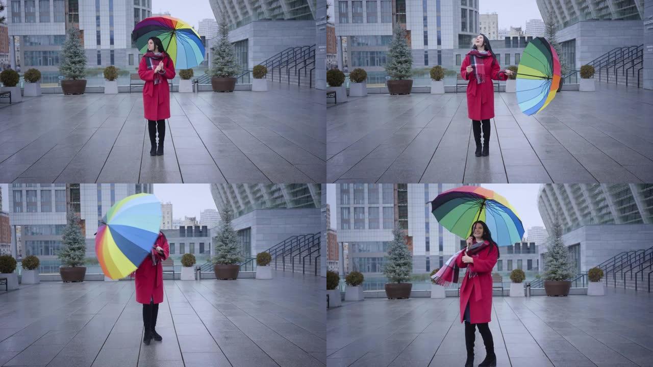 在城市街道上旋转着五颜六色伞的积极年轻白人妇女的长镜头。穿着时尚红色外套的开朗女孩微笑着面对下雨天。
