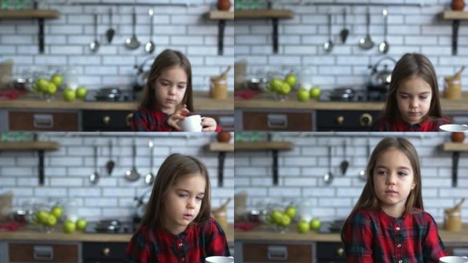 可爱的小女孩穿着方格衬衫，端着杯子坐在厨房的桌子旁