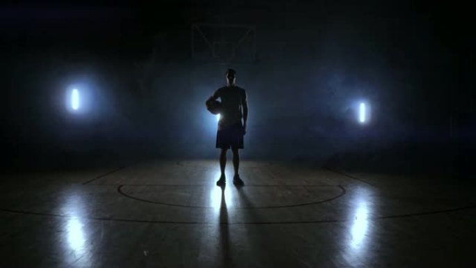 篮球运动员站在黑暗的操场上，将球握在手中，在黑暗中看着相机，慢动作的背光和烟雾笼罩着