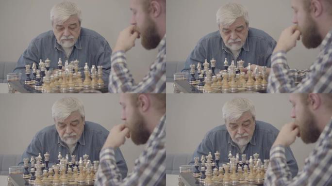 集中成熟的高加索人与成年儿子下棋的肖像。专注的老年退休人员和大胡子的年轻人在室内休息。生活方式，快乐