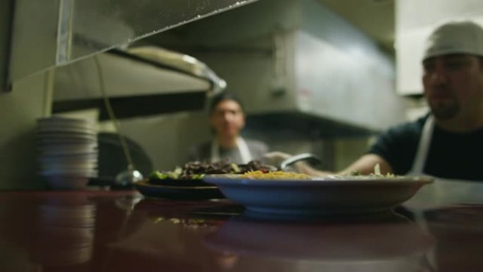 年轻的墨西哥男性厨师在一家餐厅里蒸盘子，窗户上有Fajitas和铃铛，供服务员拿起