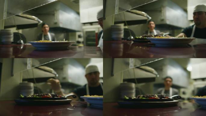 年轻的墨西哥男性厨师在一家餐厅里蒸盘子，窗户上有Fajitas和铃铛，供服务员拿起