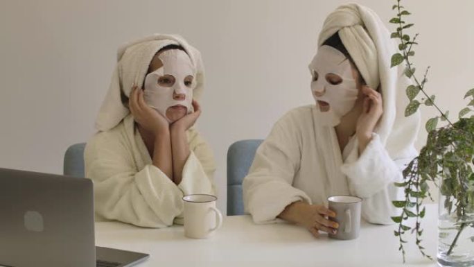 两个戴着口罩的白人成年女孩坐在桌旁聊天。穿着浴巾和浴袍在家休息的积极女性。电影院4k镜头ProRes