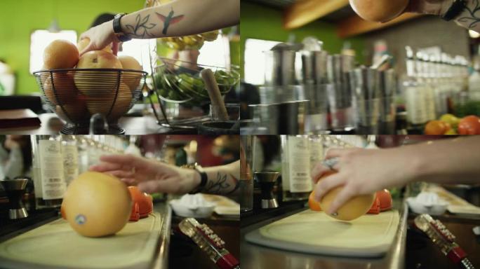 一位纹身的高加索女性调酒师从一篮子水果中拿出一个葡萄柚，将其放在砧板上，并准备在酒吧准备鸡尾酒时用刀