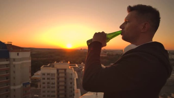 一个男人站在屋顶上喝着啤酒，沉思着世界。从高处看这座城市日落时的景色。摩天大楼的屋顶