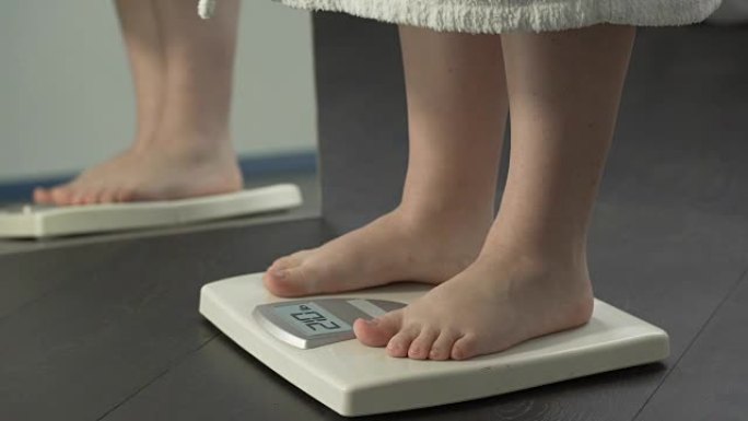 超重，穿着浴袍的胖女性踩在秤上检查饮食结果