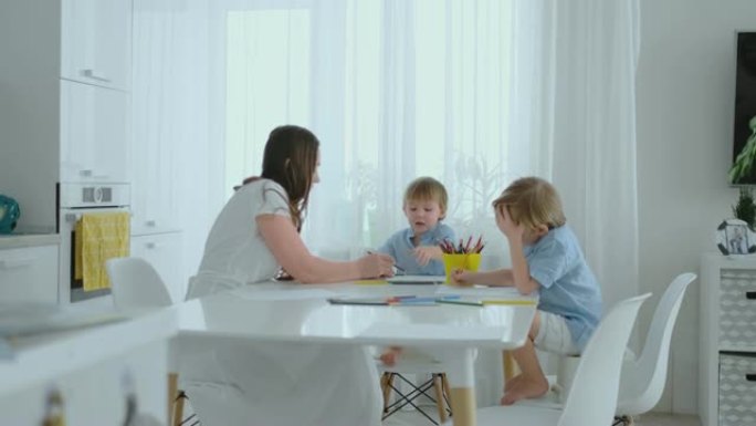 妈妈和她的两个儿子坐在厨房的桌子上，夏天在草坪上画彩色铅笔。男孩学画画，妈妈帮忙画画