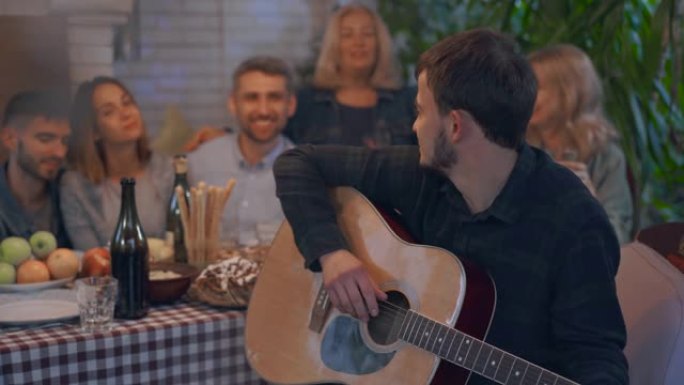一群喝酒的人，微笑着坐在后台的桌子旁。弹吉他的年轻人