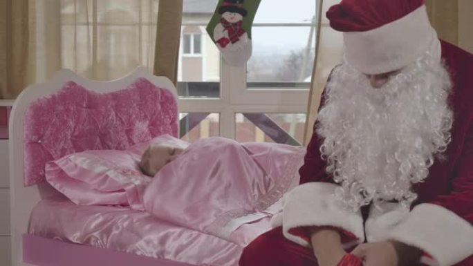 圣诞老人坐在孩子的床上，小漂亮的高加索女孩在后台醒来。孩子在家里的粉红色床上醒来，看到男人带着圣诞礼
