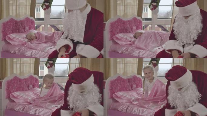 圣诞老人坐在孩子的床上，小漂亮的高加索女孩在后台醒来。孩子在家里的粉红色床上醒来，看到男人带着圣诞礼