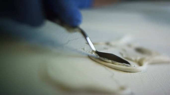 一位艺术家戴着防护手套，用调色板刀在画布上散布干墙泥/油灰的特写镜头