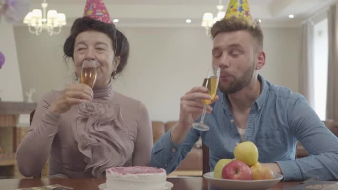 可爱的奶奶和成年的孙子戴着生日帽子在桌上碰碰眼镜