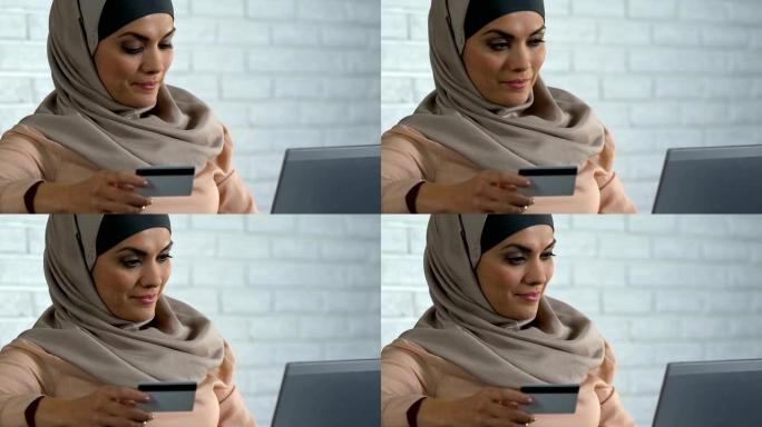 满意的穆斯林女士在笔记本电脑上插入信用卡号码，在线购物
