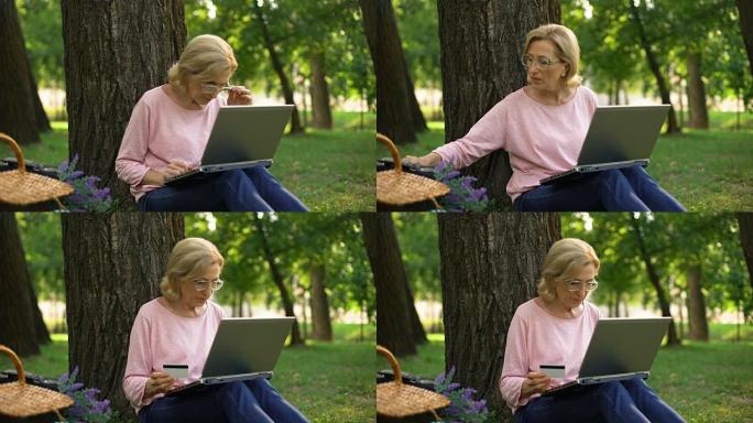 漂亮的女性养老金领取者坐在公园里，拿着笔记本电脑在网上购物