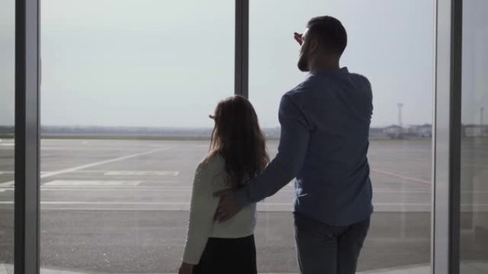 高加索男子和小女孩的轮廓的后视图，从跑道的玻璃机场门口望去。父女手握在额头上。等待飞机起飞的家庭。