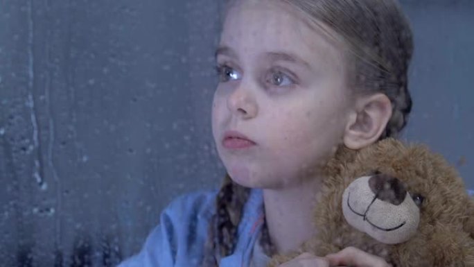孤独可爱的女孩在雨窗后面的孤儿院拥抱泰迪熊，做梦