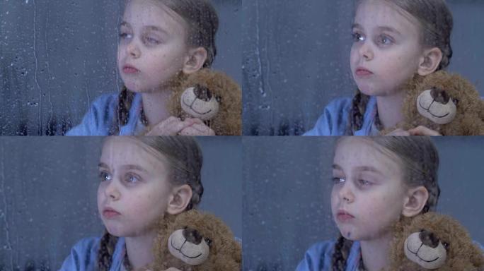 孤独可爱的女孩在雨窗后面的孤儿院拥抱泰迪熊，做梦