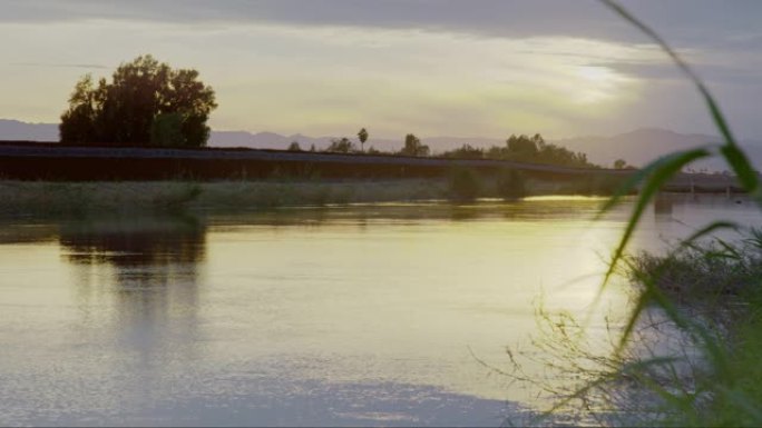 在日落时分的阴天，水沿着一条与墨西哥和美国之间的钢板边界墙 (在美国一侧) 平行的运河流下，上面是剃