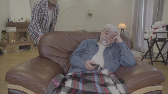 白人老人晚上在家看电视时睡着的肖像。成年儿子拿走遥控器，用毯子盖住退休人员。关怀、团结、生活方式、衰