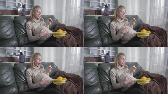 漂亮怀孕的高加索女孩吃苹果和爱抚她的肚子的肖像。她丈夫在后台的厨房里找东西。怀孕，怀孕，产妇的快乐。