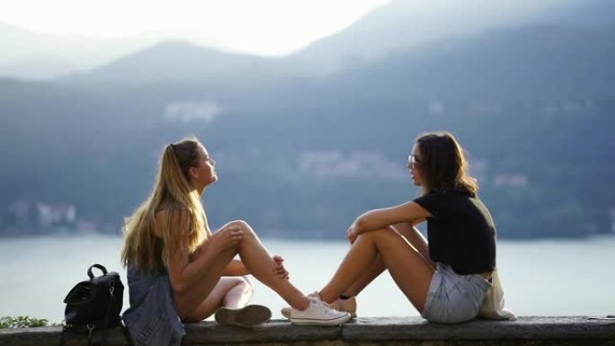 两个女孩坐在石墙上俯瞰湖泊和山脉