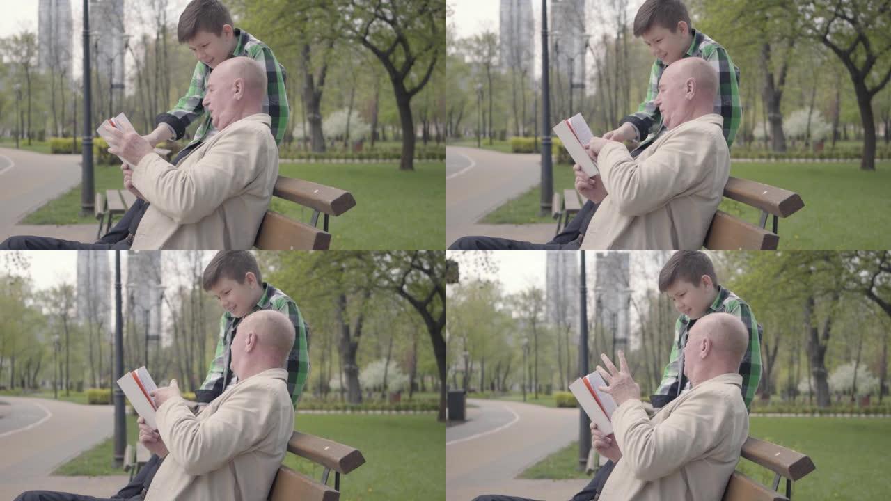 肖像可爱的爷爷和可爱的孙子坐在公园的长凳上，老人为小男孩看书。