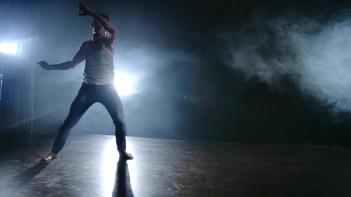现代芭蕾舞剧，一个男人从舞台上的一个坐着的位置跳起后空翻，在聚光灯下的烟雾中，在黑暗的舞台上旋转