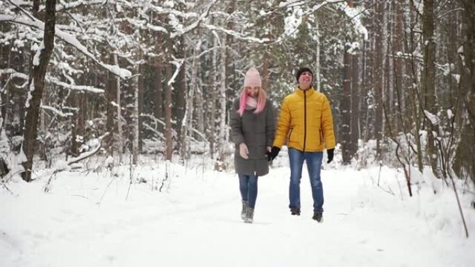 一个穿着黄色夹克的男人和一个戴着帽子和围巾的女孩在降雪期间穿过冬季森林，在圣诞节慢动作中互相笑着微笑