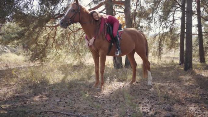 摄像机接近快乐的高加索女性马术，躺在马背上爱抚她的宠物。美丽的棕色马与年轻的黑发骑手在秋天的森林中休