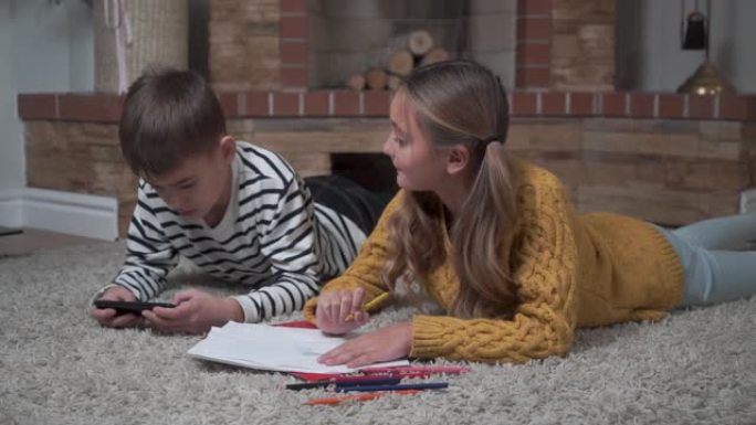 高加索可爱的女孩用蜡笔着色的特写镜头，看着她的哥哥在智能手机里玩游戏。兄弟姐妹躺在家里的柔软地毯上。