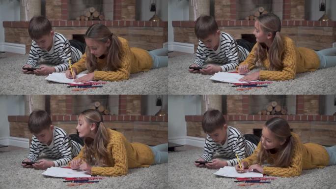 高加索可爱的女孩用蜡笔着色的特写镜头，看着她的哥哥在智能手机里玩游戏。兄弟姐妹躺在家里的柔软地毯上。