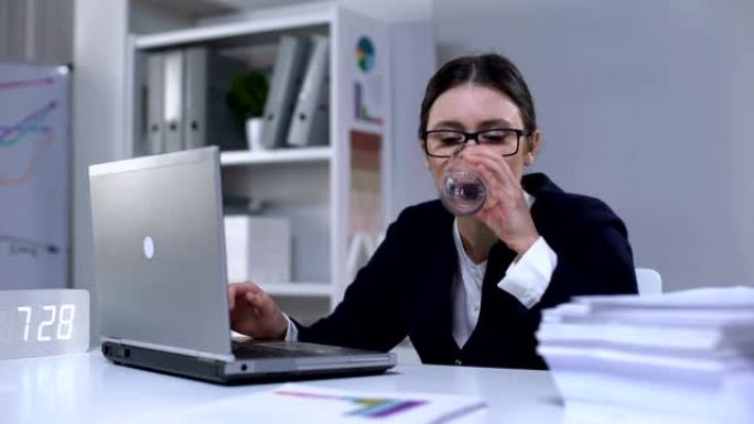 穿着西装的疲倦女人在电脑和饮用水上打字，工作到很晚
