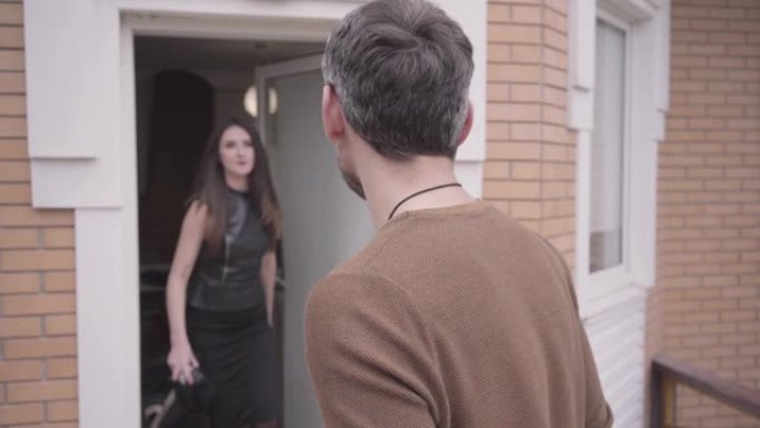一个年轻英俊的欧洲男人站在门廊前的肖像。愤怒的妻子或女友走出家门，把他的衣服扔给他。感情问题，嫉妒，