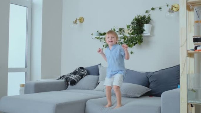 一个4岁的男孩，白色的头发和一件蓝色的衬衫在沙发上跳跃并微笑。有趣和宝贝在家里。恶作剧和纵容