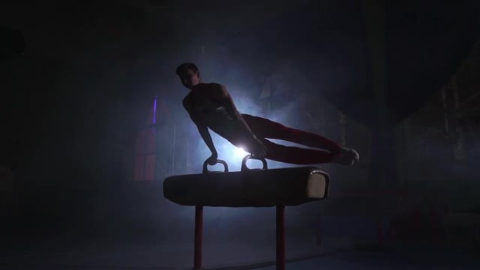 男体操运动员在黑暗的背景上表演倒立并在鞍马上旋转，并以慢动作吸烟。奥林匹克计划