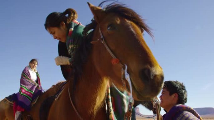 在晴朗明亮的日子里，一个十几岁的美国原住民男孩帮助一个戴着传统纳瓦霍人毯子的十几岁的女孩骑在马身上的