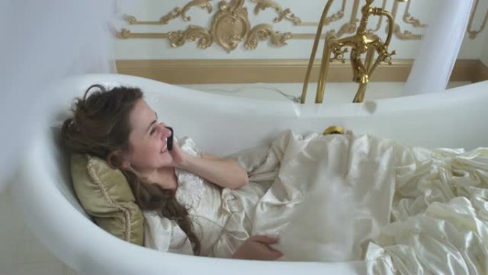 美丽的年轻女子躺在枕头上的ampty浴缸里，用手机聊天