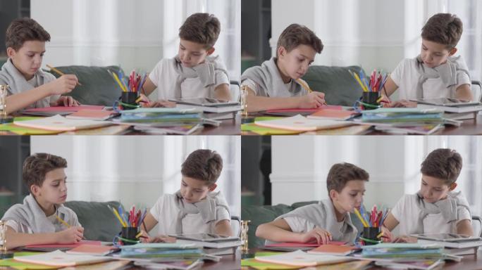 白人男生坐在桌旁做作业的肖像。黑发男孩看着他的双胞胎兄弟的作品并写下信息。兄弟姐妹在家里一起学习。