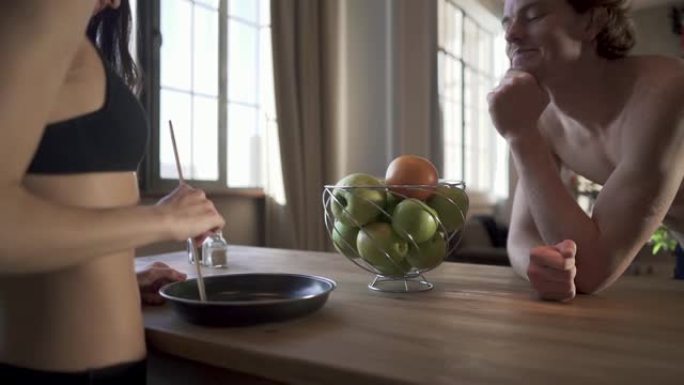 一对年轻裸男在阁楼厨房里。穿着内衣在平底锅里做饭的女孩用木勺搅动食物。