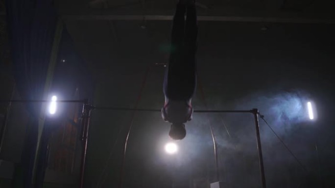 横杆上黑暗背景的体操运动员进行旋转，并以慢动作和烟雾在空中跳过自己