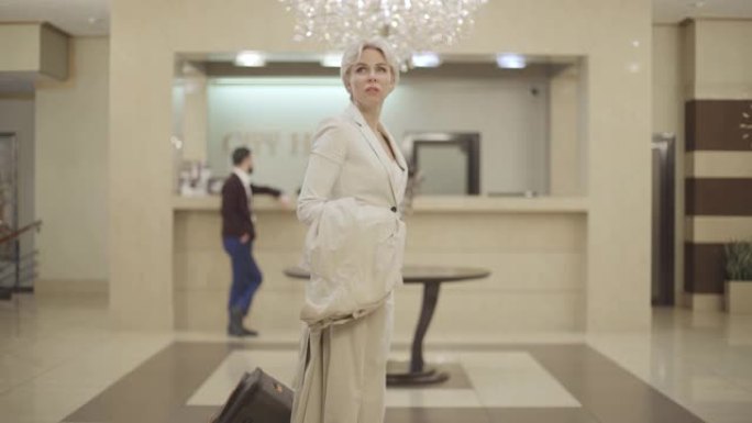 年轻的金发白人妇女的肖像在豪华的前厅环顾四周，带着旅行包离开。女商人在高级酒店路过接待处，背景模糊。