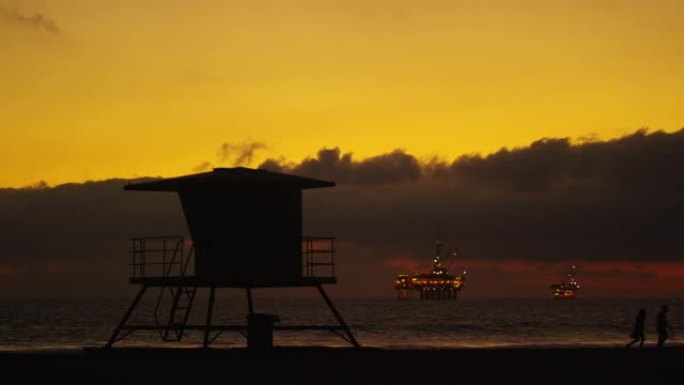 两个剪影的人在南加州亨廷顿海滩的救生员小屋旁边Walk狗，远处的地平线上有几个照亮的海上石油钻机平台