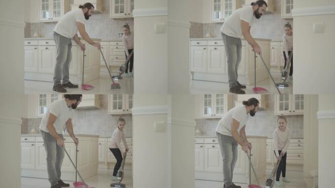 留着胡须的爸爸和可爱的小女儿在带扫帚和簸箕的新现代厨房里打扫和吸尘