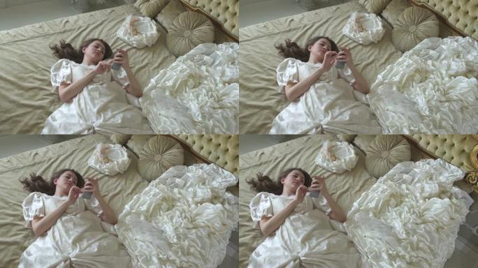 穿着舞会礼服的年轻疲惫的女人躺在金色装饰的床上，用手机发短信特写。