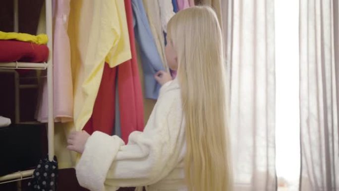 可爱的小女孩，长长的金发和灰色的眼睛来到衣柜，看着衣服，思考。美丽的孩子早上选择服装。风格，时尚，童
