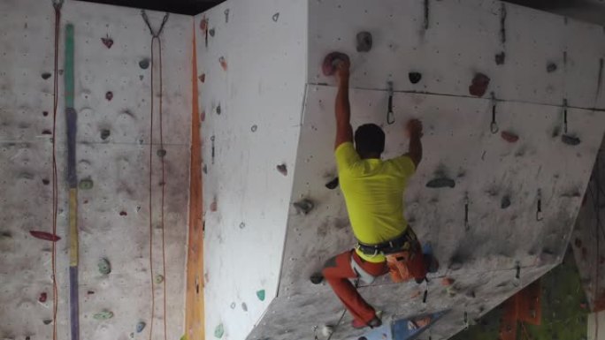年轻的攀岩者正在攀岩健身房内攀岩。苗条的漂亮男人在室内攀岩健身房的墙壁上锻炼